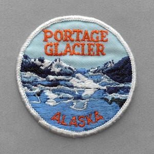 May include: A circular embroidered patch featuring a light blue sky above dark blue and white mountains and water. The text PORTAGE GLACIER is stitched in orange at the top, and the word ALASKA is stitched in orange at the bottom edge.