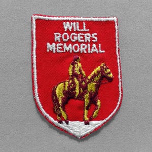 May include: A red embroidered patch with white text reading "WILL ROGERS MEMORIAL." The patch features a yellow and maroon image of a person on a horse. The patch has a white border and is set against a gray background.