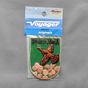 May include: A shield-shaped embroidered patch featuring a brown ruffed grouse in flight over light pink flowers on a green background. The text PENNSYLVANIA is stitched in black at the top. The patch is in a blue Voyager Originals header card.