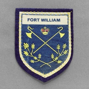 May include: A navy blue and yellow patch with the text "FORT WILLIAM". The patch features a shield design with crossed axes, a crown, and floral elements. The edges are stitched with yellow thread.