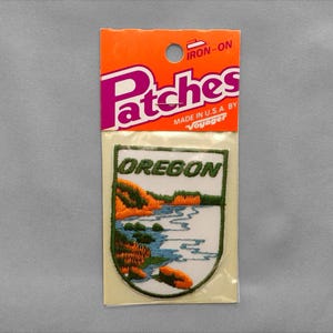 May include: An iron-on embroidered patch in original packaging labeled Patches and Voyager. The patch features a white background with the word OREGON in green letters above a landscape scene of a blue river, green trees, and orange foliage.