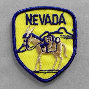 May include: A yellow and blue embroidered patch with the word "NEVADA" in blue lettering. The patch features a stylized image of a burro carrying supplies.