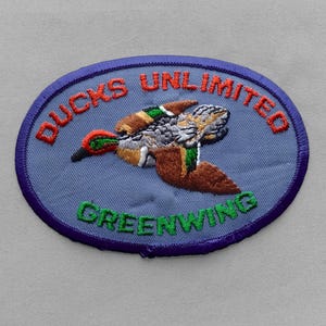 May include: An oval-shaped embroidered patch with a light blue background and a dark blue border. It features a central image of a flying duck with brown, green, and white feathers. The text DUCKS UNLIMITED is in red, and GREENWING is in green.