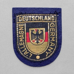 May include: A dark blue embroidered patch featuring a central shield with the German flag colors of black, red, and gold, topped by a black eagle emblem. The text DEUTSCHLAND, GERMANY, and L'ALLEMAGNE is stitched in gold thread around the shield.