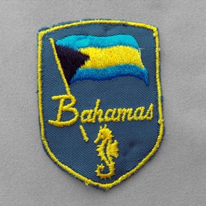 May include: A shield-shaped embroidered patch with a dark blue background and a yellow border. It features the flag of the Bahamas, the word Bahamas in yellow cursive script, and a yellow seahorse icon centered at the bottom of the patch.