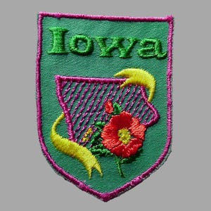 May include: Embroidered patch featuring the word "Iowa" in green, above a state outline with a floral design. The patch has a green background, a pink border, and yellow ribbon accents.