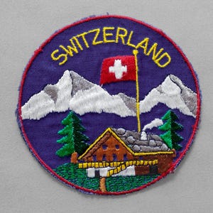 May include: A circular embroidered patch featuring a purple background with the word SWITZERLAND in yellow text. The design includes white snow-capped mountains, a red flag with a white cross, green pine trees, and a brown wooden cabin.