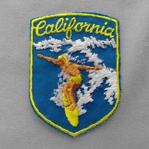 May include: A shield-shaped embroidered patch with a blue background and a yellow border. The text California is stitched in yellow at the top. A brown figure is depicted surfing on a yellow surfboard amidst white embroidered ocean waves.