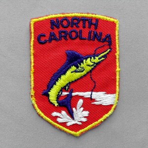 May include: A shield-shaped embroidered patch with a red background and a yellow border. It features the text NORTH CAROLINA in dark blue letters above a graphic of a jumping marlin fish in yellow and dark blue, with white water splashes.