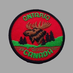 May include: Embroidered patch featuring a moose with antlers, set against a red background. The patch includes the text "ONTARIO CANADA" in green, with a black border. The design incorporates green trees and a brown moose.