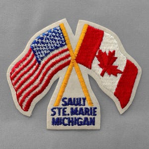 May include: Embroidered patch featuring the flags of the United States and Canada, crossed with yellow poles. The American flag has red and white stripes with blue stars. The Canadian flag has a red maple leaf. The text "SAULT STE. MARIE MICHIGAN" is below the flags.
