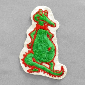 May include: Embroidered patch featuring a cartoon alligator in green with red accents. The alligator has a red bow tie and is outlined in red stitching. The patch is on a white background with a gray fabric backdrop.