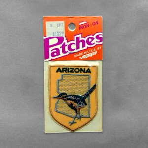 May include: An iron-on embroidered patch featuring a yellow shield shape with the word ARIZONA in black text above a black and brown roadrunner bird graphic. The patch is attached to an orange and white cardboard backing with the text Patches.