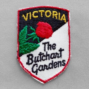 May include: Embroidered patch with a shield shape. The patch features the word "VICTORIA" in yellow, a red rose, and the text "The Butchart Gardens" in black. The patch has a red border and a black and white background.
