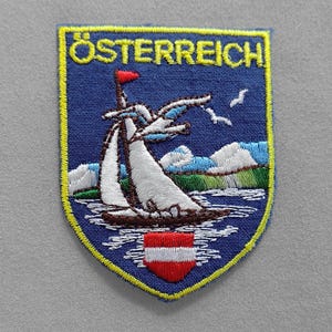 May include: Embroidered patch featuring the word "ÖSTERREICH" in yellow above a blue shield. The shield depicts a sailboat, mountains, and the Austrian flag. The patch has a yellow border.
