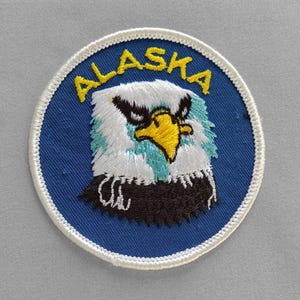 May include: Embroidered patch featuring a detailed depiction of an eagle's head. The patch is circular, with a blue background and a white border. The word "ALASKA" is arched across the top in yellow letters. The eagle has white, black, and turquoise feathers, and a yellow beak.