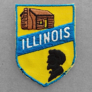 May include: A yellow and blue embroidered patch with the word "ILLINOIS" and a silhouette of a person. The patch also features a small brown cabin with a log cabin design.