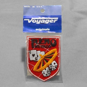 May include: A Reno, Nevada patch with a red shield design. The patch features the text "Reno Nevada" in black, along with images of playing cards, dice, and a yellow and black wheel. The patch is in a clear plastic sleeve with a blue Voyager Originals header.
