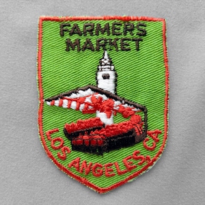 May include: A shield-shaped embroidered patch with a bright green background and a red border. It features the text FARMERS MARKET in black at the top and LOS ANGELES, CA in red at the bottom. A white tower and red structure are in the center.