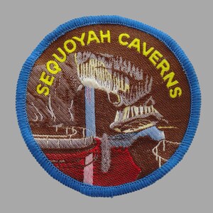 May include: Embroidered patch with a brown background and a blue border. The patch features the words "SEQUOYAH CAVERNS" in yellow, surrounding a detailed illustration of a cave with a waterfall and a red and white structure.