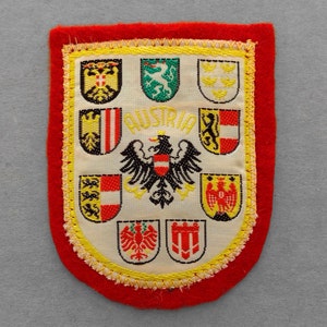 May include: A shield-shaped patch with a red border and a cream-colored interior. The patch features the word "AUSTRIA" and several colorful crests, including a black eagle. The patch is trimmed with yellow stitching.
