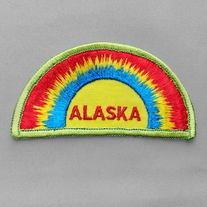 May include: Embroidered patch featuring a rainbow design with red, yellow, and blue colors. The word "ALASKA" is embroidered in red across the yellow center. The patch has a green border and is set against a gray background.
