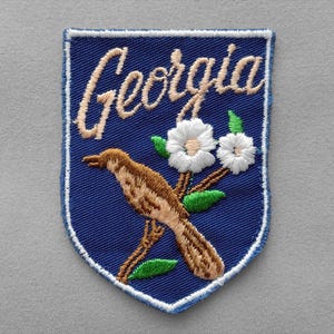 May include: A shield-shaped embroidered patch with a blue background and a white border. It features the word Georgia in tan script, a brown bird perched on a branch, and two white flowers with green leaves, all rendered in detailed thread work.
