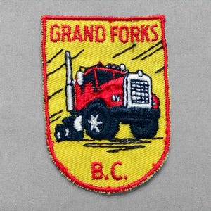 May include: Embroidered patch featuring a red and black semi-truck with a white grill and wheels. The patch has a yellow background with the words "GRAND FORKS" at the top and "B.C." at the bottom, all within a red border.