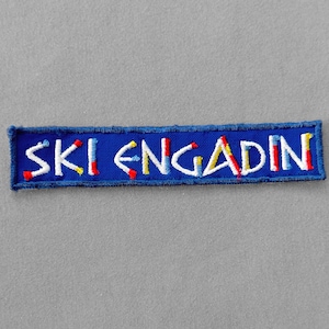 May include: A rectangular blue embroidered patch with the text "SKI ENGADIN" in white, red, and yellow. The patch has a blue border and is likely designed to be sewn onto clothing or accessories. The background is a solid gray.