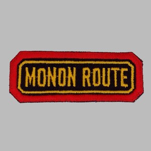 May include: Embroidered patch with the text "MONON ROUTE" in yellow letters on a black background, framed by a yellow border and a red outer border. The patch has a rectangular shape with rounded corners.