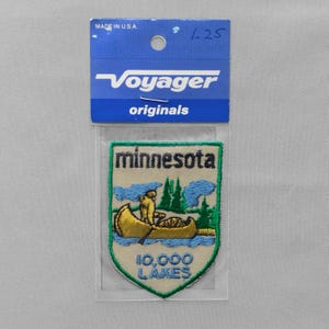 May include: A shield-shaped embroidered patch in a plastic sleeve with a blue header card labeled Voyager originals. The patch features a tan background, a gold canoe with a person paddling, green pine trees, blue water, and the text Minnesota 10,000 Lakes.