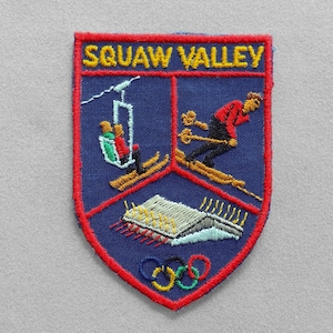 May include: Embroidered patch with the words "SQUAW VALLEY" in yellow. The patch features a skier, a ski lift, a building, and Olympic rings. The patch is blue with a red border.