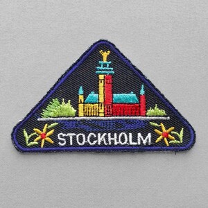 May include: A triangular embroidered patch with a dark blue background featuring a colorful depiction of a building with a tower, green foliage, and yellow flowers. The word STOCKHOLM is stitched in white capital letters at the bottom center.