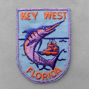 May include: A rectangular embroidered patch with a light blue background featuring a purple marlin fish leaping over an orange boat. The text KEY WEST is embroidered in orange at the top, and the word FLORIDA is embroidered in orange at the bottom.