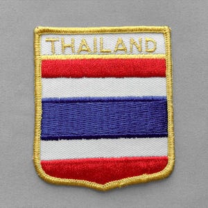May include: A rectangular embroidered patch with rounded bottom corners featuring the flag of Thailand. The design includes horizontal red, white, and blue stripes with the word THAILAND stitched in gold thread across a white top border.