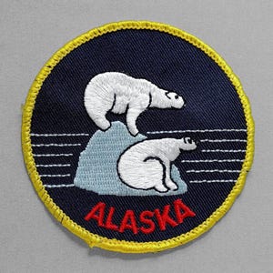 May include: Embroidered patch featuring two white polar bears on a light blue iceberg against a dark blue background. The word "ALASKA" is stitched in red at the bottom, with a yellow border. The patch is round.