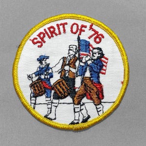 May include: A circular embroidered patch with a yellow border on a white background. It features three figures in colonial attire playing drums and a fife, with a flag. The text SPIRIT OF '76 is arched in red thread across the top of the patch.