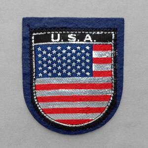 May include: A shield-shaped embroidered patch featuring a dark blue border and a central design of the United States flag. The top black bar displays the text U.S.A. in white letters. The flag design includes red and silver stripes and white stars.
