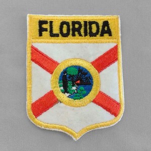May include: Embroidered patch featuring the word "FLORIDA" in black on a yellow banner. The patch has a white background with a red diagonal cross and a circular emblem with a tree and water scene.