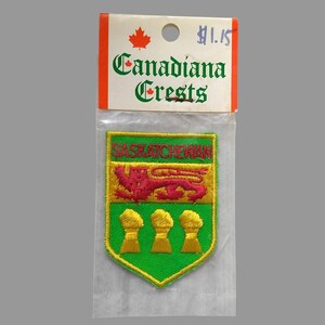 May include: A green and gold embroidered patch with the coat of arms of Saskatchewan. The patch is on a white background with a price tag that says "Canadiana Crests $1.15".