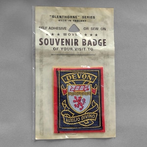 May include: A woven souvenir badge from Devon, England, in a clear plastic package. The badge features a crest with the word "DEVON" in gold, a red lion, and the phrase "AUXILIO DIVINO". The badge is self-adhesive or sew-on.