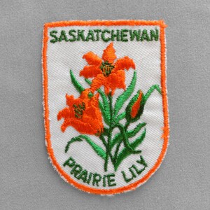 May include: Embroidered patch featuring a white background with an orange border. The patch displays an orange prairie lily with green leaves and the text "SASKATCHEWAN" and "PRAIRIE LILY" in green.