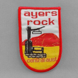 May include: A red, white, and yellow embroidered patch with the text "ayers rock central aust" and a black and white illustration of a train.