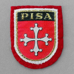 May include: A red and white embroidered patch with the word "PISA" in gold letters. The patch features a white cross on a red background, framed by gold and white trim, and a red border.