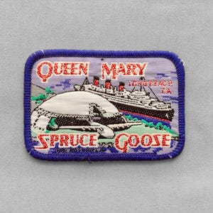 May include: A rectangular embroidered patch with a blue border featuring the Queen Mary ship and the Spruce Goose aircraft. The text reads Queen Mary, Long Beach CA, and Spruce Goose in red lettering against a light purple and blue background.