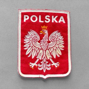 May include: A rectangular fabric patch featuring a red background with a white embroidered eagle emblem centered below the white text POLSKA. The eagle has a gold crown and gold claws, and the entire patch is bordered by a white stitched edge.