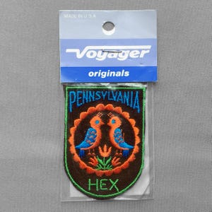 May include: A shield-shaped embroidered patch in a clear plastic package with a header card labeled Voyager originals. The patch features a dark brown background, two orange and blue birds, a central orange flower, and the text Pennsylvania Hex.