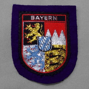 May include: A purple felt patch with a white embroidered coat of arms. The coat of arms features a lion, a mountain range, and a blue and white checkered pattern. The text "BAYERN" is above the coat of arms.