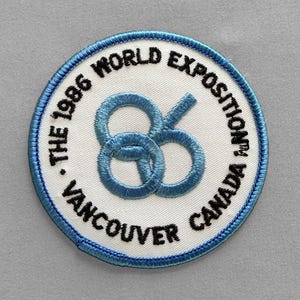 May include: Embroidered patch featuring the text "The 1986 World Exposition Vancouver Canada" in black, surrounding a blue intertwined "86" design on a white background, with a blue border.