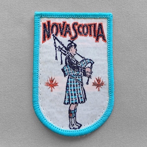 May include: A white patch with a light blue border featuring the word "Nova Scotia" in red and a drawing of a bagpiper in a kilt. Two red maple leaves are on either side of the bagpiper.